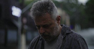 Orta yaşlı bir adam, ciddi bir şekilde aşağıya bakıyor, yağmurda sırılsıklam, hüzünlü bir yansıma ve duygusal ağırlık ifade ediyor.
