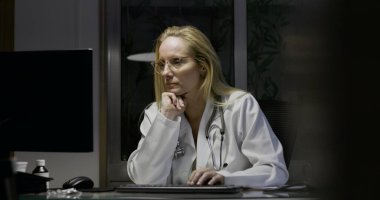 Kadın doktor gece vardiyasında çalışıyor, bilgisayar ekranını izlerken çenesini dinlendiriyor, derin düşüncelere dalmış ve modern klinik setin tıbbi öğeleriyle çevrili.