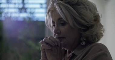 Gözleri kapalı ve elleri sımsıkı bağlı yaşlı bir kadın sessizce dua ediyor ya da pencerenin yanında duygusal bir yansıma yaşıyor.