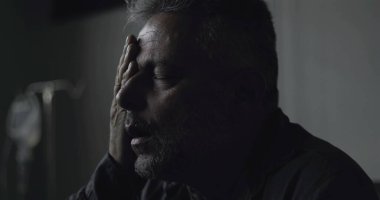 Klinikte gözleri kapalı oturan orta yaşlı bir adamın yakın plan çekimi, tıbbi belirsizlik anında sessizce ağrıyı ve yorgunluğu işliyor.