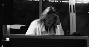 Yalnızlık içinde yanmış kadın doktor, gözleri kapalı ve elleri boynunda, zihinsel yorgunluk ve duygusal çöküntüyü ifade eden gri ölçekli iş yerinde