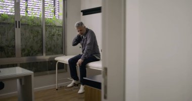 Sıkıntılı bir adam tıbbi odada tek başına, muayene masasında oturmuş, boyun tutarak, sağlık kontrolünden sonra rahatsızlık ya da duygusal yorgunluk belirtileri gösteriyor..