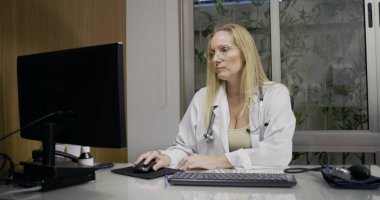 Tıp bürosunda masaüstü bilgisayarı üzerinde çalışan, hasta veri girişine odaklanan ya da klinik çalışma oturumunda rapor yazan kadın doktor