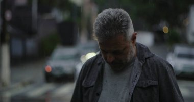 Orta yaşlı bir adam yağmurun altında duruyor. Islak giysiler ve kasvetli bir ifade. Etrafı bulanık arabalar ve kasvetli sokak atmosferiyle çevrili.