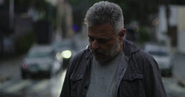 Orta yaşlı bir adam yağmurun altında duruyor. Islak giysiler ve kasvetli bir ifade, bulanık arabalar ve karamsar sokak atmosferiyle çevrili.
