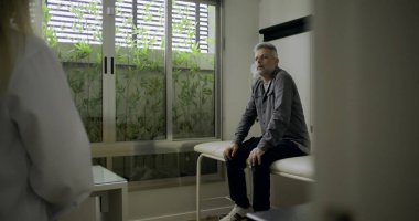 Modern tıp bürosunda muayene masasında oturan erkek hastayla konuşurken not alan kadın doktor, tıbbi danışma süreci ve özenli ifade ile devam ediyor.