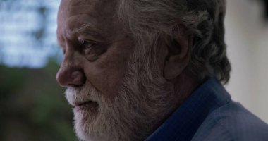 Yaşlı adam nazikçe gülümserken yumuşak arka plan mavisi ve kapalı mekan ortamında nazik ve düşünceli bir ifadeyle yan yana bakıyor.