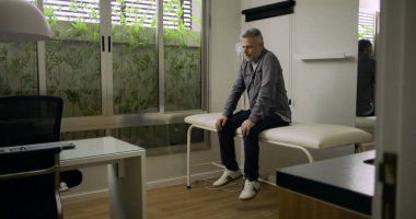 Muayenehanede muayene masasında tek başına oturan orta yaşlı bir adam. Muayeneden sonra düşünceli ve sıkıntılı görünüyor. Klinik kaplıcada sakin bir yansıma anı.