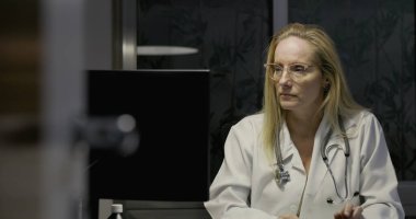 Kadın doktor bilgisayar ekranına odaklanmış, geceleri klinikte klavyeye yazı yazıyor, reçeteli şişeler ve hasta belgelerine bakılıyor.