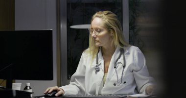 Masada geç saatlere kadar çalışırken not alan kadın doktor, tıbbi malzemeler ve belgelerle çevrili, odaklanmış ifadeli bilgisayar monitöründen uzağa bakıyor.