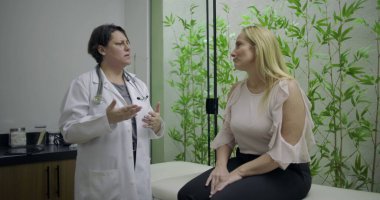 Doktor, yeşil bambu bitkisi geçmişi olan klinikteki sağlık danışmanlığı sırasında muayene masasında oturan orta yaşlı kadın hastayla konuşuyor.