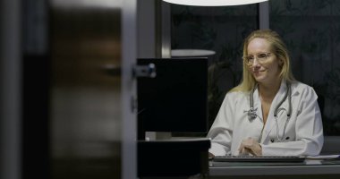 Geç saatlere kadar çalışan bayan doktor bilgisayar monitöründeki bilgileri gözden geçiriyor, elinde ilaç şişesi, modern tıp ofisinde düşünceli ve ciddi bir ifade.