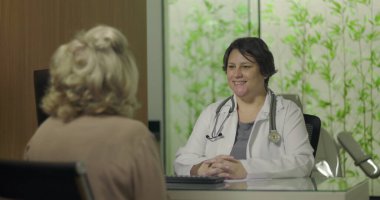 Steteskoplu gülümseyen doktor profesyonel klinikteki tıbbi danışmanlık sırasında yaşlı kadın hastayla samimi bir şekilde temas kuruyor.