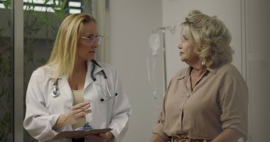 Klinik ziyareti sırasında yaşlı kadın hastaya teşhisi açıklayan kadın doktor, hem ayakta duruyor hem de arkadaki ekipmanlarla konuşuyor.