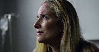 Klinikteki kadının yoğun, duygusal bir ifadeyle yan tarafa baktığı yakın plan. Arkaplanda tıbbi tedavi veya iyileşme öneren serum var.