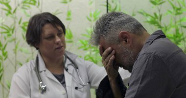 Sıkıntılı orta yaşlı erkek hasta yüzünü kapatırken, kadın doktor klinikte destek sunuyor, zor tıbbi haberlere duygusal ifadeyle tepki veriyor.