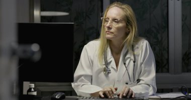 Kadın doktor bilgisayar ekranına odaklanmış, geceleri klinikte klavyeye yazı yazıyor, reçeteli şişeler ve hasta belgelerine bakılıyor.