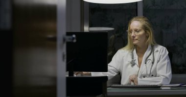 Geç saatlere kadar çalışan bayan doktor bilgisayar monitöründeki bilgileri gözden geçiriyor, elinde ilaç şişesi, modern tıp ofisinde düşünceli ve ciddi bir ifade.