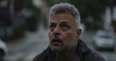 Yukarı bakan orta yaşlı bir adam, yağmurda ellerini kaldırmış, duygusal sorgulama ya da ruhani bir katman.