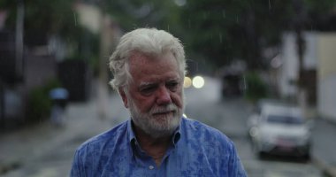 Yaşlı bir adam yağmurun altında üzgün gözlerle ve duygusal ifadeyle, dudakları sımsıkı ve kasvetli bir şekilde duruyor.