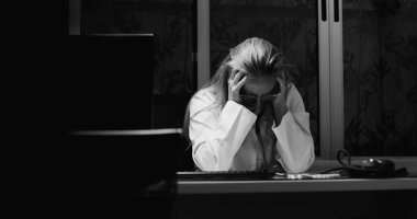 Masasında oturan kadın doktor, elleri ve gözleri dramatik bir şekilde siyah ve beyaz kareyle kapatılmış bir şekilde, tıbbi klinikte geç saatlere kadar yatıyor.