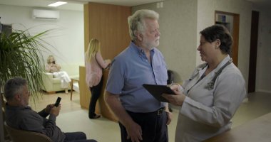 Üst düzey bir adam, klinikteki kadın doktorla dijital tablet hakkındaki bilgileri gözden geçiriyor. Koridorda sağlık hizmetleri ve yakınlardaki hastalarla konuşuyor.