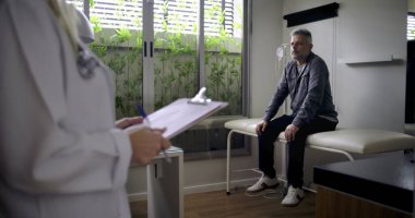 Orta yaşlı erkek hasta muayene odasında muayene masasında beklerken, kadın doktor ön planda yazı yazıyor.