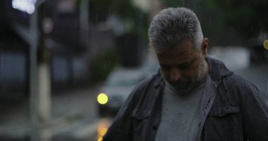 Orta yaşlı bir adam yağmurun altında duruyor. Islak giysiler ve kasvetli bir ifade, bulanık arabalar ve karamsar sokak atmosferiyle çevrili.