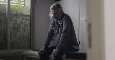 Klinik odasında tek başına oturan, arkasında serum olan, sessizce aşağıya bakan, tıbbi konsültasyondan ya da teşhisten sonra duygusal olarak ağırlığını koyan bir adam.