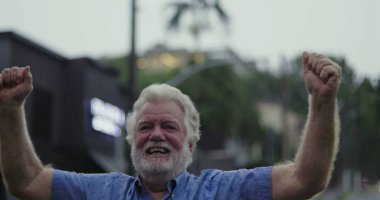 Mavi gömlekli yaşlı adam kollarını kaldırdı ve neşeli bir ifade takındı. Yağmurun altında coşkuyla gülümsüyordu.