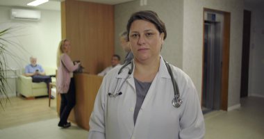 Kadın doktor, klinik koridorundaki kameraya bakıyor. Steteskop ve laboratuvar önlüğü giyiyor. Arkaplanda sakin ve dikkatli bir ifade var.