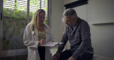 Hasta muayene masasında oturan orta yaşlı bir erkeğe danışmanlık yaparken elinde dosya tutan kadın doktor.