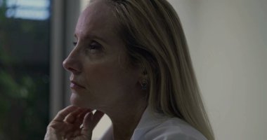 Yan profildeki kadın doktor, elleri çenesinin altında, gözleri klinik ortamda derin bir yansıma anında ileri bakıyor.