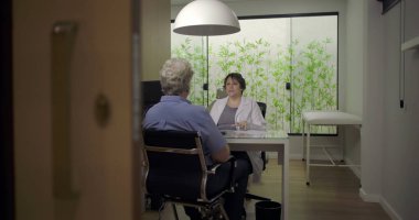 Klinikteki özel danışma sırasında yaşlı erkek hastayla konuşan doktor, profesyonel sağlık hizmetleri interaktifi hakkında yakın perspektif için kapı aralığından çerçevelenmiş