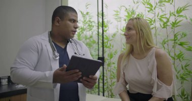 Parlak klinikte dijital tablet kullanan orta yaşlı kadın hastaya danışmanlık yapan erkek doktor, açık iletişimi teşvik ediyor ve kişiselleştirilmiş tıbbi bakım