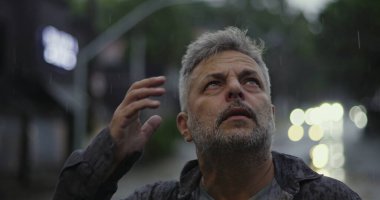 Orta yaşlı bir adam yağmurda dalgın dalgın bir ifadeyle yukarı bakıyor, boş şehir sahasında öylece duruyor.