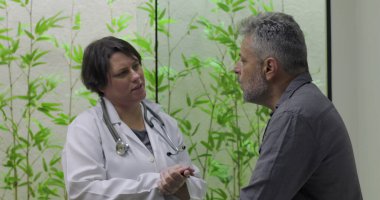 Orta yaşlı erkek hasta modern klinikte tıbbi danışmanlık sırasında teşhis veya tedavi seçeneklerini açıklayan kadın doktoru dikkatle dinliyor.