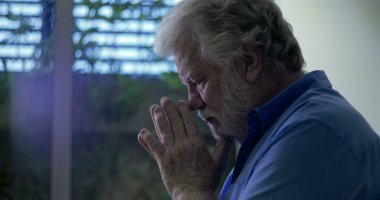 Yüzü nazik bir ifadeyle yukarı bakan yaşlı bir adam dua veya yansıma, elleri yavaşça birbirine yapışmış pencerenin yanında duruyor