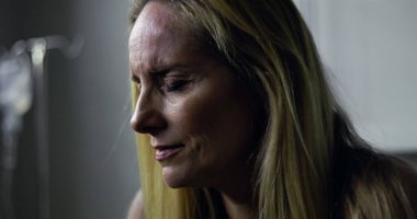 Klinikteki kadının yoğun, duygusal bir ifadeyle yan tarafa baktığı yakın plan. Arkaplanda tıbbi tedavi veya iyileşme öneren serum var.