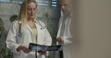 Kadın doktor önplanda güvenle dururken, erkek meslektaşı tıbbi danışma sırasında arka plandaki röntgeni inceliyor.