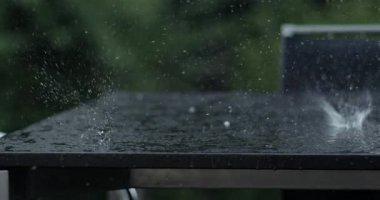 Yağmurun masa yüzeyine vurması ve yavaş çekimde damlacıklar halinde patlaması, arka planda yeşil yapraklar, sıçrama ve çarpmanın detaylı görüntüsü