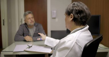 Doktor, tıp bürosunda kağıt ve cam masadaki belgelerle baş başa görüşmede orta yaşlı bir hastaya hitap ediyor.