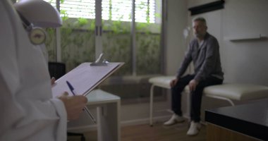 Orta yaşlı erkek hasta klinik muayene odasında otururken, elinde steteskoplu bir pano tutan kadın doktor.