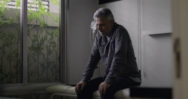 Klinik odasında tek başına oturan, arkasında serum olan, sessizce aşağıya bakan, tıbbi konsültasyondan ya da teşhisten sonra duygusal olarak ağırlığını koyan bir adam.