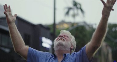 Kolları geniş ve yüzü göğe yükselmiş yaşlı bir adam, ruhsal rahatlama, minnettarlık veya duygusal teslimiyet ifadesiyle yağmuru kucaklıyor.