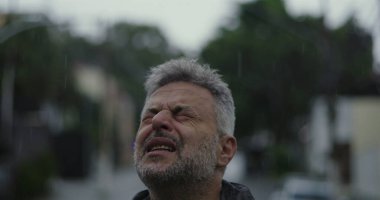 Orta yaşlı adam yağmurda ellerini kaldırarak yukarı bakıyor. Duygusal sorgulama ifadesi.