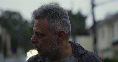 Orta yaşlı bir adam endişeli bir ifadeyle, gergin duruşu ve şehir sokaklarında duygusal ağırlığıyla yan yana bakıyor.