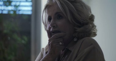 Yakın profil içinde orta yaşlı bir kadın, sakin, yansıtıcı bir ifadeyle, yumuşak arka plan ışığıyla çerçevelenmiş bir şekilde ileriye bakıyor.