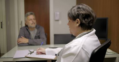 Doktor, tıp bürosunda kağıt ve cam masadaki belgelerle baş başa görüşmede orta yaşlı bir hastaya hitap ediyor.