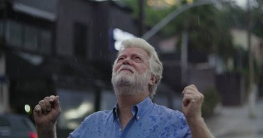 Yağmurda ayakta duran yaşlı adam, elleri açık ve duygusal ifade ile yukarı bakıyor keder, kafa karışıklığı, ya da ruhsal özlem şehir caddesinde